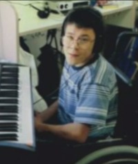 Photo of Nicholas Dean, a boy with dark hair and light-brown skin, sitting in a wheelchair in front of an electric keyboard. He is wearing glasses and a striped shirt.