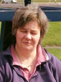 Photo of Sharon Greenop, a middle-aged woman with short brown hair, photographed outdoors. The camera seems to have caught her off guard, since she has her eyes closed mid-blink.