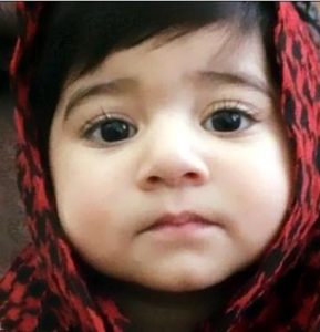 Photo of Inaya Ahmed. She is a toddler girl with light skin and dark brown hair and eyes. She is wearing a red and black checked headscarf.