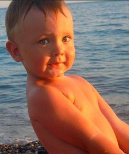 Photo of Misha Kordyukova, a baby with fair skin and short blond hair, photographed against a background of water.