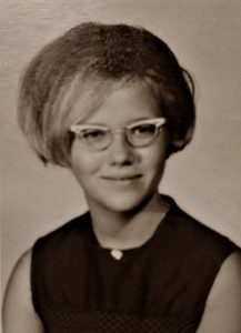 Sepia portrait photo of Sharon McCleary as a young woman. She has fair skin and hair cut in a bob; she is wearing old-fashioned glasses and a black sleeveless top.