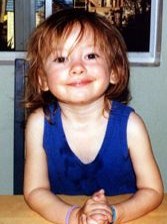 Photo of Eliza Jane Scovill, a toddler with tangled auburn hair, fair skin, and brown eyes wearing a blue dress. She is smiling for the camera.