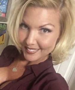 Selfie-style photo of Kathleen West, a woman with fair, slightly tanned and freckled skin and blonde hair. She is wearing a maroon blouse left open to show a little cleavage, and a heart-shaped gold necklace.