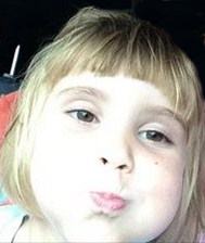 Photo of Olivia Cart, a little girl with blonde hair in bangs, very pale skin and brown eyes. She is pursing her lips and puffing her cheeks. The photo is taken from very close in, and is overexposed.