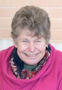 Photo of Helen Dansie, an older woman with fair skin and short curly gray hair. She is wearing a flowered scarf with a pink sweater and smiling broadly.