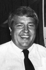 Black and white photo of Ed Dossett, a middle-aged, heavyset white man wearing a shirt and tie. He is smiling.