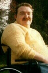 Photo of Dominic McCarthy. He is a large, middle-aged man wearing a sweater and sitting in a wheelchair. He has short brown hair, fair skin, and a mustache.