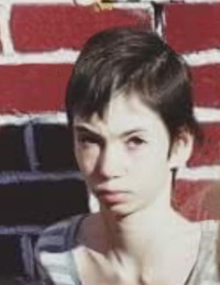 Photo of Sabrina Ray. She is a thin girl with pale skin and dark brown hair in a boy cut. Her expression is solemn.
