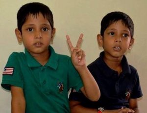 Photo of Sarthak and Varad, twin boys. They have black hair and light-brown skin, and are wearing collared shirts, one green and one blue. The boy in the green shirt is holding up two fingers in a "V" sign.