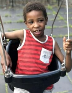 Boy with brown skin and short curly brown hair, smiling, seated in an adapted swing and gripping the chains the swing hangs by. He is wearing a red striped tank top.
