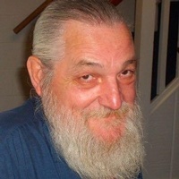 Photo of a man with a full gray beard and mustache, ruddy skin, and neatly combed back gray hair. He is wearing a blue shirt; his expression is amused.