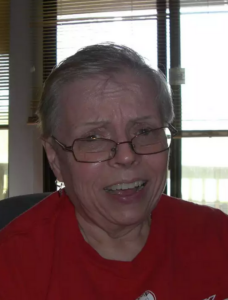 Photo of an older woman wearing a red shirt and glasses, which are slipping down her nose. She has light skin, and her short gray hair is wispy.