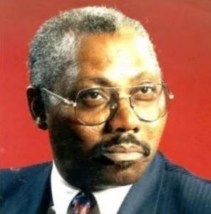 Portrait photo of a man in a suit, wearing glasses. He has brown skin and a solemn expression. His short curly hair is graying.