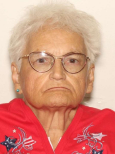 Photo of an older woman with wispy white hair, tan skin, cat-eye glasses, and a red shirt printed with stars. She is scowling at the camera, corners of her lips pulled down.