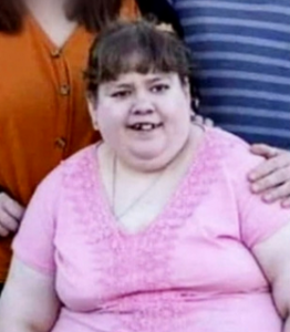 Photo of a young woman, heavyset with pale skin and brown hair, smiling. She is wearing a pink blouse.