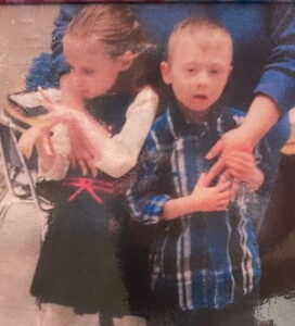 Photo of two small children being held by an adult off-frame. The child on the left is a little girl with her dark-blond hair pulled away off her forehead, looking off to the left. She has pale skin. Her hands are blurred from flapping them. She is wearing a black dress with a white blouse. On the right is a little boy with wispy blond hair and pale skin, wearing a dark blue flannel shirt and hugging the adult's hand to his chest. His mouth is slightly open in a neutral expression.