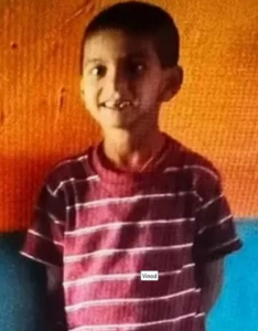 Photo of a boy with tan skin, short black hair, and brown eyes, smiling for the camera. He has a gap in his front teeth and is wearing a red striped shirt.