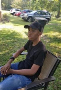 Photo of a young Black teenage boy, wearing jeans, black T-shirt, and baseball cap, sitting in a lawn chair. In the background is a neatly mown lawn and some parked cars.