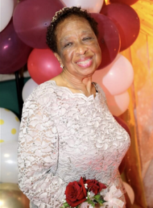 Photo of an older woman in a lacy white dress, wearing a corsage of red roses on her wriest. She is smiling. Her skin is brown, and her short black hair is curly. There are balloons in the background.