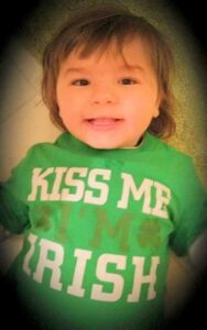 Photo of a toddler with fine, straight brown hair, dark-brown eyes, and light skin, smiling. He is wearing a green T-shirt that reads, Kiss me, I'm Irish.