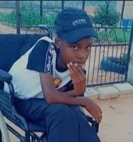 Photo of a boy sitting in a wheelchair. He is wearing a black and white T-shirt and black baseball cap, and has his chin propped on the fingers of one hand. His skin is brown and his eyes are dark-brown.