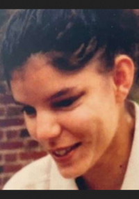 Photo of a woman with light skin and dark hair, smiling and looking down to the left. She is wearing a white shirt. There is a brick wall in the background.