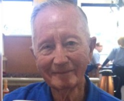 Photo of an older man with light skin, white hair, a receding hairline, large ears, and a friendly expression. He is wearing a blue collared shirt.