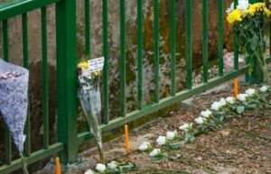 Flowers left at a green railing in front of a shallow canal. There are three bouquets of yellow flowers and a line of white roses.