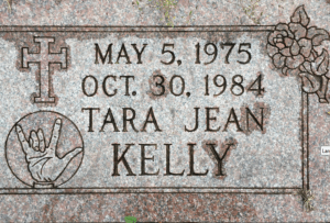 Tombstone for Tara Jean Kelly, May 5 1975 to October 30 1984. It is decorated with a cross and a hand signing I Love You in ASL.