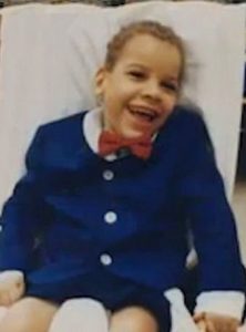 Photo of Trevor Horn, a little boy with olive skin and short curly brown hair. He is smiling. He is wearing a blue suit with a red bow tie that is a bit too big for him.