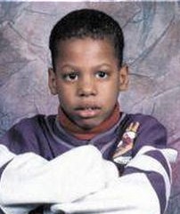 School photo of a boy with short, very curly black hair and brown skin and eyes. He is wearing a loose sweater, and has his arms folded in front of him. His expression is neutral.