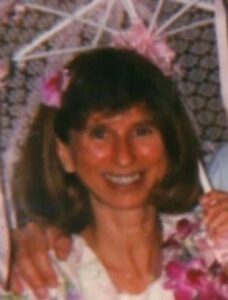 Photo of Cynthia Hrisco, a middle-aged woman with shoulder-length brown hair and bangs, holding a flowered parasol and wearing a flowered dress.