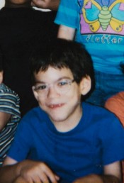 Photo: A boy with pale skin and a mop of dark brown hair, wearing large glasses and a blue sweater, smiling for the camera.
