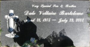 Gravestone reading, "Very special son & brother, Dale Voltaire Bartolome, April 28, 1975 to July 29, 2002." The dark granite stone is decorated with a carved cross with a rosary hanging from it.