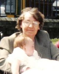 Photo of Evelyn Joel. She is a middle-aged woman with pale skin and graying dark-brown hair and glasses, holding a baby.