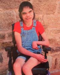 Photo of Kate Bugmy. She is a young woman sitting in a wheelchair, with her dark hair tied back. She is wearing overalls and an orange T-shirt. Her limbs are very thin.