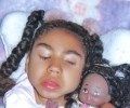 Photo of a girl with light-brown skin, her dark-brown hair done in two braids. She is leaning back against a pillow with her eyes closed; she is wearing light makeup. Next to her on the pillow is a doll with matching light-brown skin and dark-brown hair. 