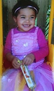 Photo of Tanilla Warrick-Deaves. She is a toddler with tan skin and curly black hair. She is wearing a pink dress, and holding a ribbon in her hands.