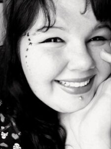 Black and white photo of a woman with dark hair and pale skin. Her make-up is neatly done; there are tiny stars painted along her cheek and a piercing in her lower lip. She is smiling.
