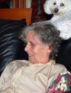Woman with short, curly gray hair, face in profile; she has light skin. She is wearing a beige silk blouse. On the back of the couch she is sitting on is a small, fluffy white dog.
