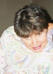 Photo of Mindy Pace, a young woman with short brown hair. She is wrapped in a paisley blanket, with her head bent forward, smiling and peeking at the camera through her bangs.