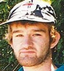 Photo of a man with fair skin and dark-blond hair, wearing a black and white print baseball cap. He has a stubbly beard and mustache.