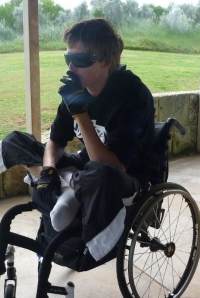 Photo of Tyron Honeywood, a young man with brown hair and light skin sitting cross-legged in a wheelchair and smoking a cigarette. He is wearing sunglasses, gloves, and black athletic pants and hoodie.