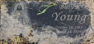 Photo of a gravestone partly buried in sandy dirt. It reads "Billy R. Young, October 18, 1961 to March 11, 2014," and is etched with an image of Jesus with hands outstretched.