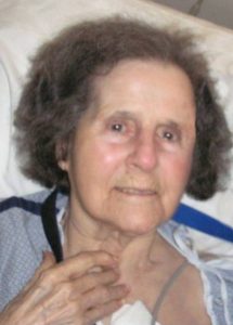 Photo of an elderly woman with gray hair and pale skin, lying back against a pillow and smiling slightly. She is wearing a hospital gown.