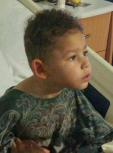 Photo of Tyrael McFall, a young boy with medium-brown skin and curly black hair. He is sitting in a hospital bed. He has mild microcephaly. He is wearing a paisley shirt.