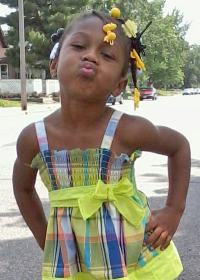 Photo of Kendrea Johnson. She is an African-American girl wearing a yellow dress, her hair in hair clips. She has her hands on her hips and is leaning forward, lips puckered, as though she is trying to kiss the camera.