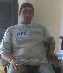 Photo of a middle-aged man with brown skin, a black durag on his head, wearing a loose gray T-shirt and jeans. 