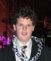 Photo of a teenage boy with fair skin and brown, curly hair, wearing a black suit and a lei draped around his neck.