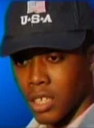 Photo of a teenage boy with brown skin, wearing a black baseball cap embroidered with an American flag and the legend USA. His expression is neutral.